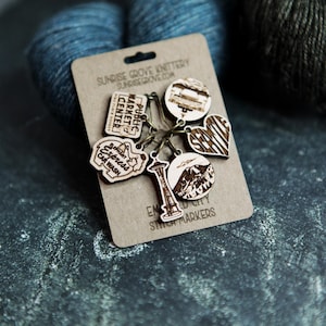 May include: A set of six wooden stitch markers featuring Seattle landmarks, including the Space Needle, Pike Place Market, and the Seattle skyline. The markers are labeled "Emerald City Stitch Markers" and are made by Sunrise Grove Knittery.