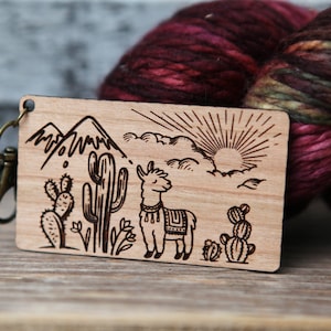 May include: A wooden llama keychain with a mountain, cactus, and sun design. The keychain is brown and has a brass key ring.