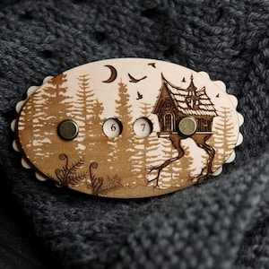 May include: Wooden brooch with a forest scene, featuring a house on stilts, trees, birds, and a crescent moon. The brooch has three metal fasteners and the numbers 6 and 7. The brooch is set against a gray knitted background.