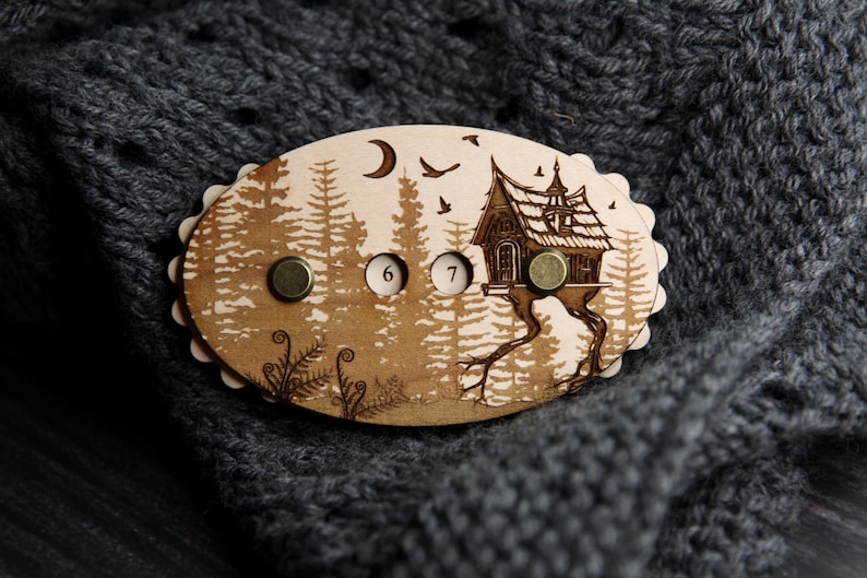 May include: Wooden brooch with a forest scene, featuring a house on stilts, trees, birds, and a crescent moon. The brooch has three metal fasteners and the numbers 6 and 7. The brooch is set against a gray knitted background.
