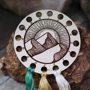 May include: A wooden thread organizer with 16 holes, featuring a laser-engraved design of a mountain range with a sun rising behind it. The organizer has three strands of embroidery floss in green, gold, and white.