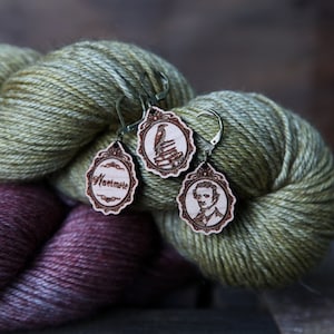 May include: A pair of wooden earrings with a vintage design. One earring features a raven perched on a stack of books, the other features a portrait of Edgar Allan Poe. The earrings are hanging on a skein of green and brown yarn.