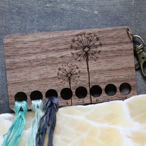 May include: Wooden thread organizer with a dandelion design and eight numbered slots. The organizer has a metal clip for attaching to a bag or keychain.  Four strands of embroidery floss are threaded through the slots.
