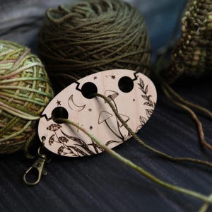 May include: A wooden yarn swift with a laser-etched design of mushrooms, stars, and a crescent moon. The swift is connected to green and brown yarn balls. A metal clasp is attached to the swift. The yarn is in shades of green and brown.