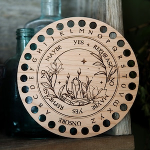 May include: A wooden circular divination board with a crystal and floral design. The board has a circle of letters around the edge and the words "Yes", "Rephrase", "No", and "Maybe" in the center.