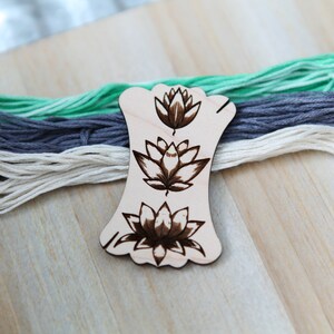 May include: A wooden thread holder with three lotus flower designs burned into the surface. The holder is on a wooden surface with three skeins of yarn in the background.