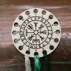 May include: A wooden thread organizer with a circular design featuring runes and numbers. The organizer has multiple holes for holding threads, with three different colored threads visible. The background is a copper-colored surface.