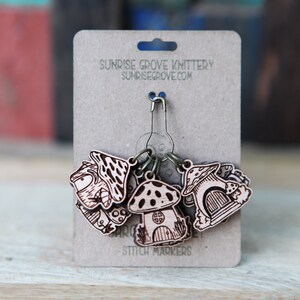 May include: Set of six wooden mushroom stitch markers with a metal ring and a safety pin. The markers are decorated with a mushroom design and the text "Sunrise Grove Knittery Sunrisegrove.com Stitch Markers".