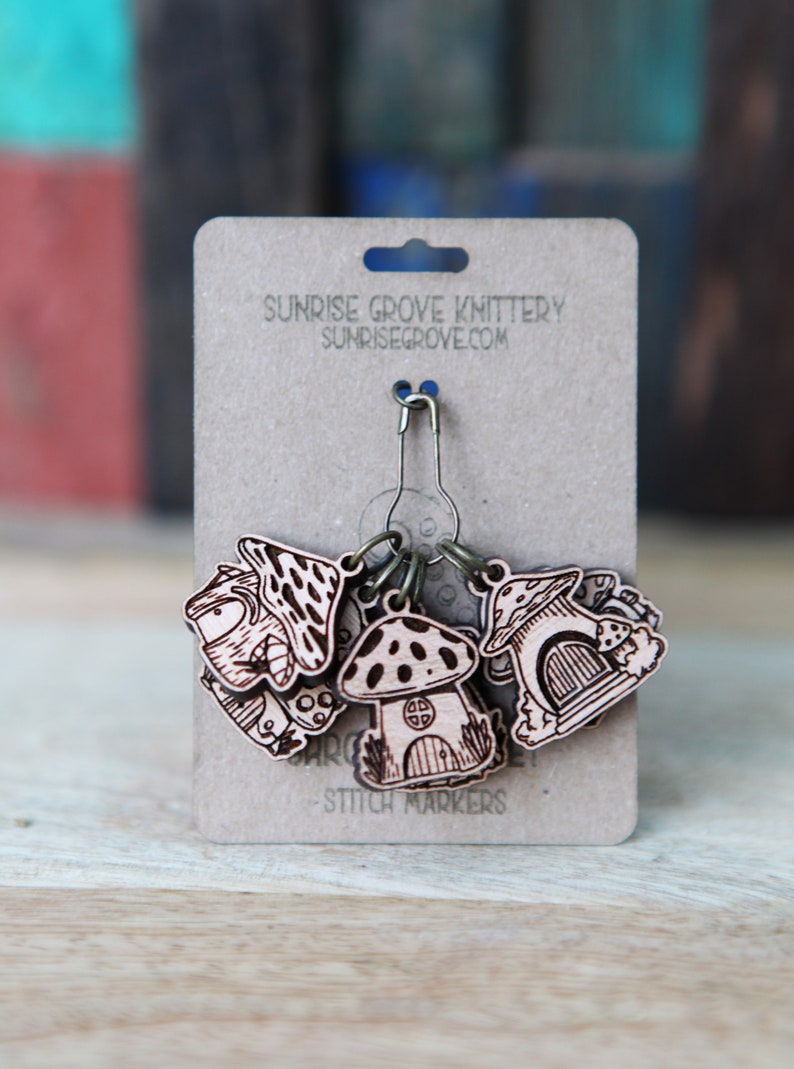 May include: Set of six wooden mushroom stitch markers with a metal ring and a safety pin. The markers are decorated with a mushroom design and the text "Sunrise Grove Knittery Sunrisegrove.com Stitch Markers".