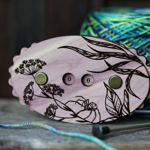 May include: A wooden yarn bowl with a floral design and a spider. The bowl has three counters with the number "0" on each. The bowl is sitting on a wooden surface next to a ball of yarn.