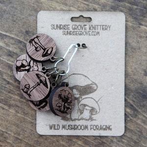 May include: Set of six wooden mushroom stitch markers with a silver metal ring. The markers are engraved with different types of mushrooms and the text "WILD MUSHROOM FORAGING". The card also includes the text "SUNRISE GROVE KNITTERY SUNRISGROVE.COM".