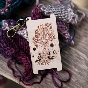 May include: A wooden stitch marker with a laser-etched design of a tree with celestial symbols, including a moon, stars, and a sun. The marker is attached to a metal ring.