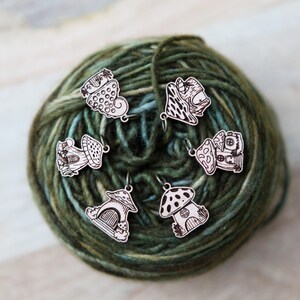 May include: Six wooden mushroom stitch markers with different designs, including a heart, a house, and a mushroom with polka dots. The markers are on a ball of green yarn.