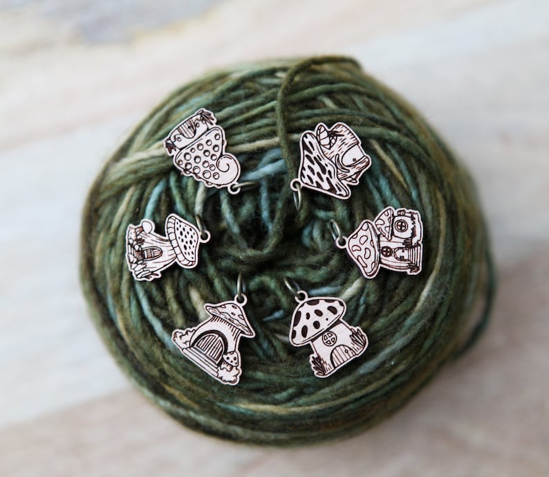 May include: Six wooden mushroom stitch markers with different designs, including a heart, a house, and a mushroom with polka dots. The markers are on a ball of green yarn.