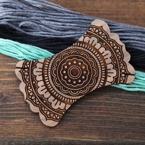 May include: Wooden thread holder with a mandala design. The holder is brown and has two slots for holding thread. The holder is sitting on a wooden surface with three skeins of thread in gray, black, and teal.