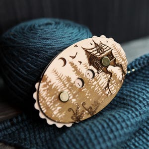 May include: A wooden yarn swift with a forest and house design, featuring a teal yarn ball and knitted fabric. The swift has a light brown finish with a detailed illustration of a house on a tree, birds, and a crescent moon.