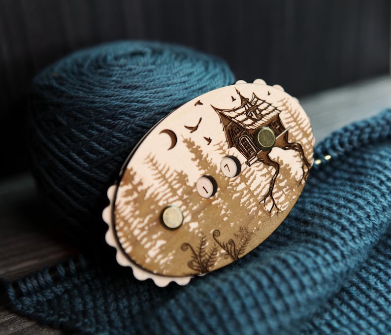 May include: A wooden yarn swift with a forest and house design, featuring a teal yarn ball and knitted fabric. The swift has a light brown finish with a detailed illustration of a house on a tree, birds, and a crescent moon.