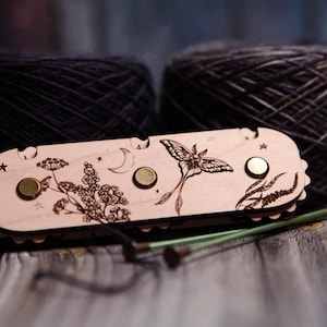 May include: A wooden knitting needle gauge with a laser-etched moth and floral design, featuring a crescent moon and stars. Two dark gray yarn balls are in the background. Knitting needles are visible.