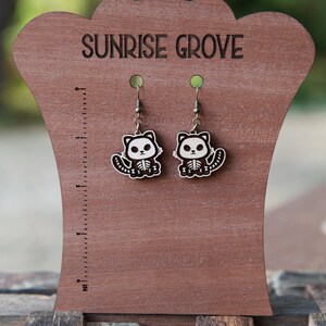May include: A pair of wooden cat skeleton earrings on a brown wooden display stand. The stand has the words "Sunrise Grove" engraved on it.
