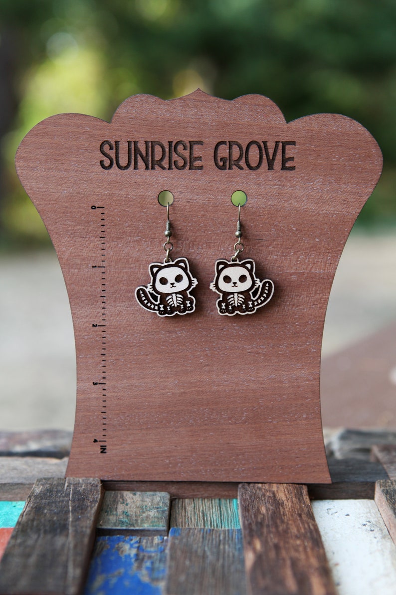 May include: A pair of wooden cat skeleton earrings on a brown wooden display stand. The stand has the words "Sunrise Grove" engraved on it.