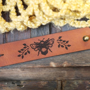 May include: A brown leather bracelet with a black engraved bee and floral design. The bracelet has a brass button closure.