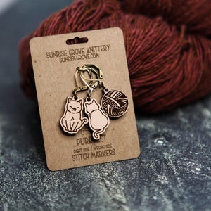May include: A set of wooden stitch markers on a brown card with the text "PURRfect" and "Sunrise Grove Knittery". The markers feature cat and yarn ball designs. A ball of red yarn is in the background.