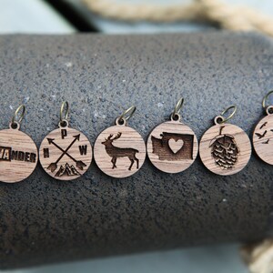 May include: Six wooden round charms with different designs, including a state outline with the word "WANDER", a compass with arrows, a deer, a state outline with a heart, a hop cone, and a bird on a branch with flying birds.