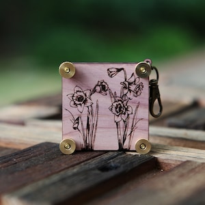 May include: A square wooden keychain with a laser-etched daffodil design. The keychain has brass-colored hardware and a clasp. The wood has a light pinkish hue, and the daffodils are depicted in black lines.