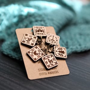 May include: A collection of six square wooden stitch markers with engraved folk art designs, including birds, deer, and trees. The markers are attached to a brown card with the text "STRIKE MAKE STITCH MARKERS". The background is a teal knitted fabric.