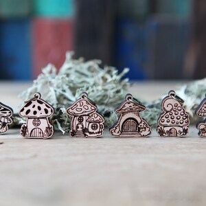 May include: Six wooden mushroom house charms with intricate details. Each charm features a different design with a mushroom cap and a small house inside. The charms are made of light brown wood and have a rustic, whimsical style.