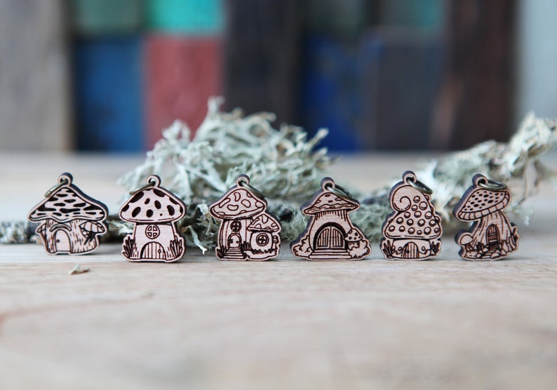 May include: Six wooden mushroom house charms with intricate details. Each charm features a different design with a mushroom cap and a small house inside. The charms are made of light brown wood and have a rustic, whimsical style.