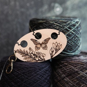 May include: A wooden yarn holder with a butterfly and floral design, resting on top of three skeins of yarn. The holder has three holes and a metal clasp. The yarn is various shades of blue, green, and brown.