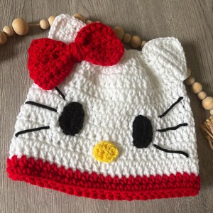 May include: A white crochet hat with a red bow, black eyes, yellow nose, and red trim. The hat is designed to resemble a cartoon cat. The hat is handmade.