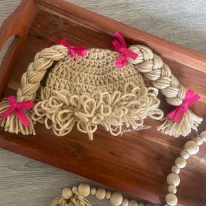 May include: A crocheted baby hat in tan yarn, designed to resemble a doll's hair. The hat features two braided yarn pigtails and a fringe of yarn around the face. Bright pink bows accent the braids. The text "yarn creation" is visible.