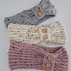 Può includere: Tre fasce per capelli lavorate a maglia in grigio, crema e malva. Ogni fascia presenta un motivo a maglia strutturato ed è adornata da due bottoni in legno. Le fasce sono esposte su una superficie bianca.