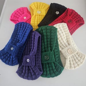 May include: Assortment of colorful knitted headbands with different button embellishments. The headbands are made of a soft, textured yarn and are available in a variety of colors, including yellow, pink, red, blue, green, and white.