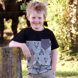 Puede incluir: Un niño pequeño con una camiseta gris y negra con un estampado de caras de animales. La camiseta tiene un bolsillo gris en el lado izquierdo. El niño sonríe y mira a la cámara.