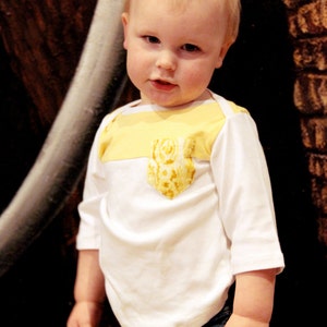 Peut inclure: Un jeune enfant portant une chemise blanche à manches longues avec une poche à motif jaune et or et une bordure jaune autour du cou. L'enfant porte un jean bleu.