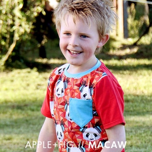 Puede incluir: Un niño pequeño con una camisa roja de manga corta con estampado de panda y un bolsillo azul. La camisa tiene un patrón geométrico con colores rojo, naranja y blanco. El texto "APPLE+FIG // MACAW" es visible en la parte inferior de la camisa.