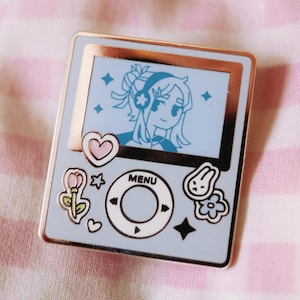 May include: A light blue enamel pin shaped like a retro music player. The screen displays a cartoon girl with headphones. The pin has a gold border and features various cute icons, including hearts, flowers, and stars. The word "MENU" is also visible.
