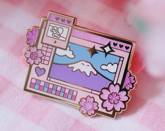 Può includere: Uno spillo in smalto dorato che raffigura uno schermo di computer con un paesaggio montano, nuvole e stelle. Lo schermo ha un bordo rosa e una tavolozza di colori rosa e blu. Lo spillo è decorato con fiori di ciliegio rosa.