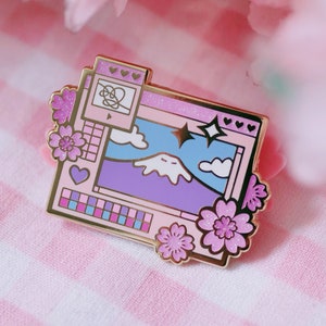 May include: A gold-toned enamel pin depicting a computer screen with a mountain landscape, clouds, and stars. The screen has a pink border and a pink and blue colour palette. The pin is decorated with pink cherry blossoms.