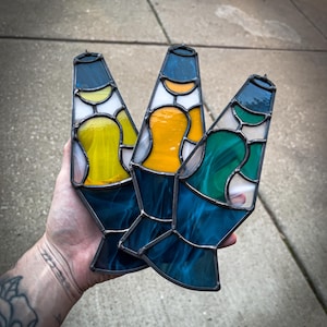 May include: Three stained glass lava lamp designs in shades of blue, yellow, orange, green, and white. Each piece has a dark metal frame and a unique color combination. The lamps are held up against a gray concrete background.
