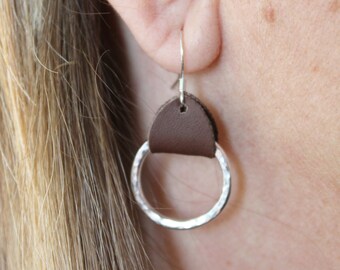 May include: Silver hoop earrings with a brown leather accent. The hoop earrings have a hammered texture.
