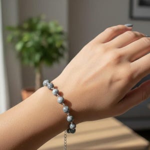 May include: A beaded bracelet with gray and white stones is displayed on a wrist. The bracelet is made of small, round beads strung together with a silver chain. The hand has gray nail polish.