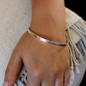 May include: A silver bangle bracelet with a hammered texture. The bracelet is on a person's wrist.
