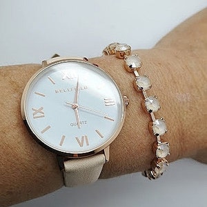 Op de afbeelding: Een roségouden horloge met een witte wijzerplaat en Romeinse cijfers voor de uren. Het horloge heeft een beige leren band en wordt gecombineerd met een roségouden armband met heldere kristallen.