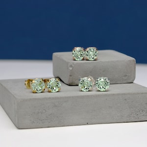 May include: Three pairs of gold stud earrings with light green round gemstones. The earrings are displayed on two grey concrete platforms.