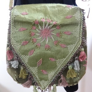 Puede incluir: Un bolso bandolera de terciopelo verde con bordado floral rosa y ribete de cuentas. El bolso tiene borlas en la parte inferior y una correa larga.
