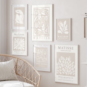 Puede incluir: Un conjunto de seis impresiones de arte enmarcadas en una gama de colores beige neutro. Las impresiones presentan diseños abstractos en blanco sobre un fondo beige. Las impresiones están dispuestas en una pared blanca con una unidad de estantería blanca y una silla de mimbre con un cojín blanco.